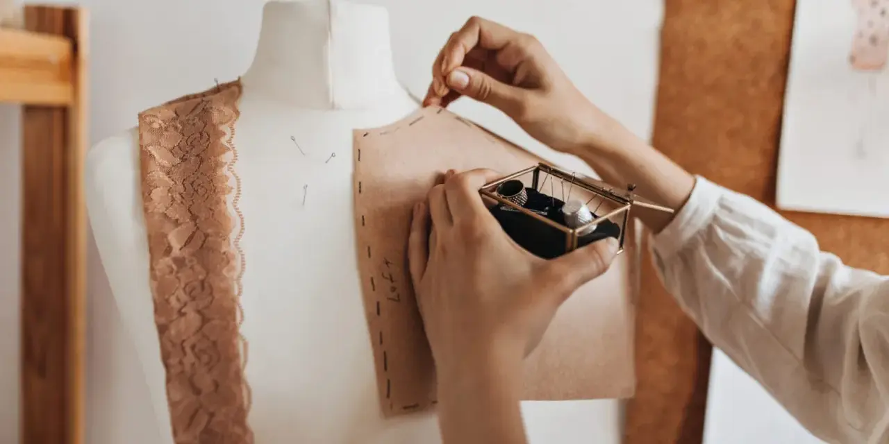 Mani che lavorano su un manichino, creando un modello di cartone. Un corso di modellistica in pieno svolgimento, con pizzi e accessori da cucito a portata di mano.