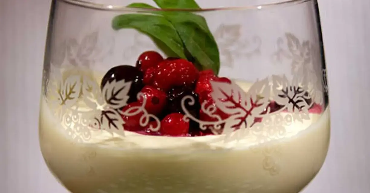 En elegant glas med vit chokladmousse, toppad med färska bär och ett grönt blad. Glaset har en vacker dekor av löv.