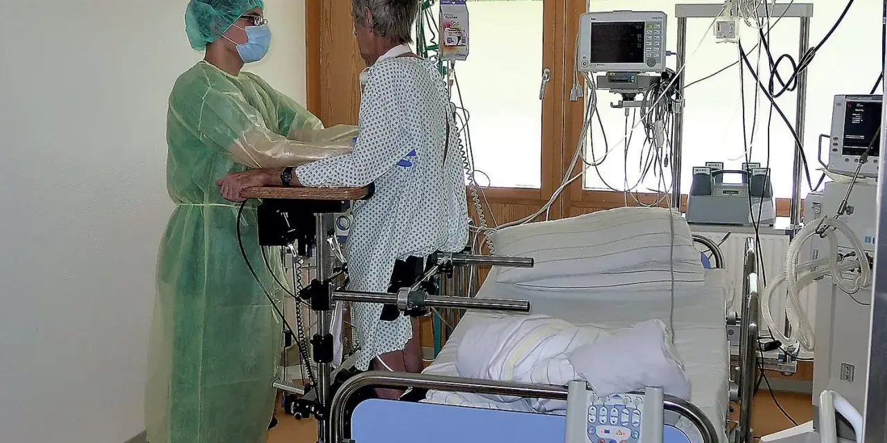 Pflegekraft unterstützt Patientin bei der Reha nach Intensivstation. Sie üben das Aufstehen.