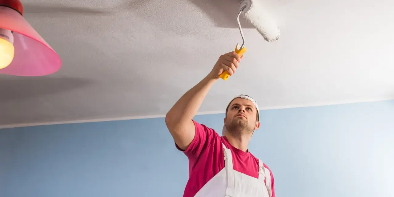 Un homme en salopette blanche et t-shirt rose utilise un rouleau pour peindre un plafond. Il est concentré sur sa tâche, montrant comment peindre un plafond.