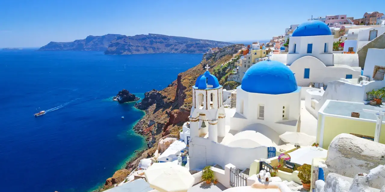 Santorini wycieczki: białe domy z niebieskimi kopułami na klifie nad błękitnym morzem. Statek płynie w oddali.