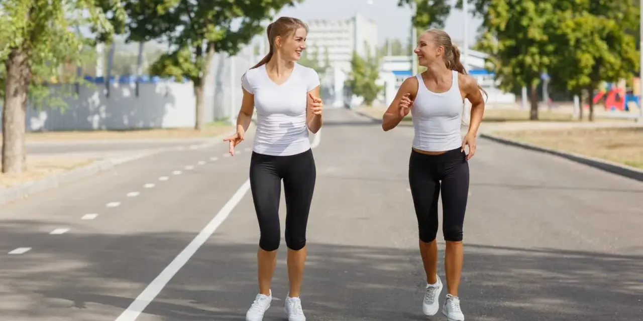 Due donne sorridono mentre praticano diversi tipi di camminata sportiva su una strada asfaltata, con alberi e edifici sullo sfondo.