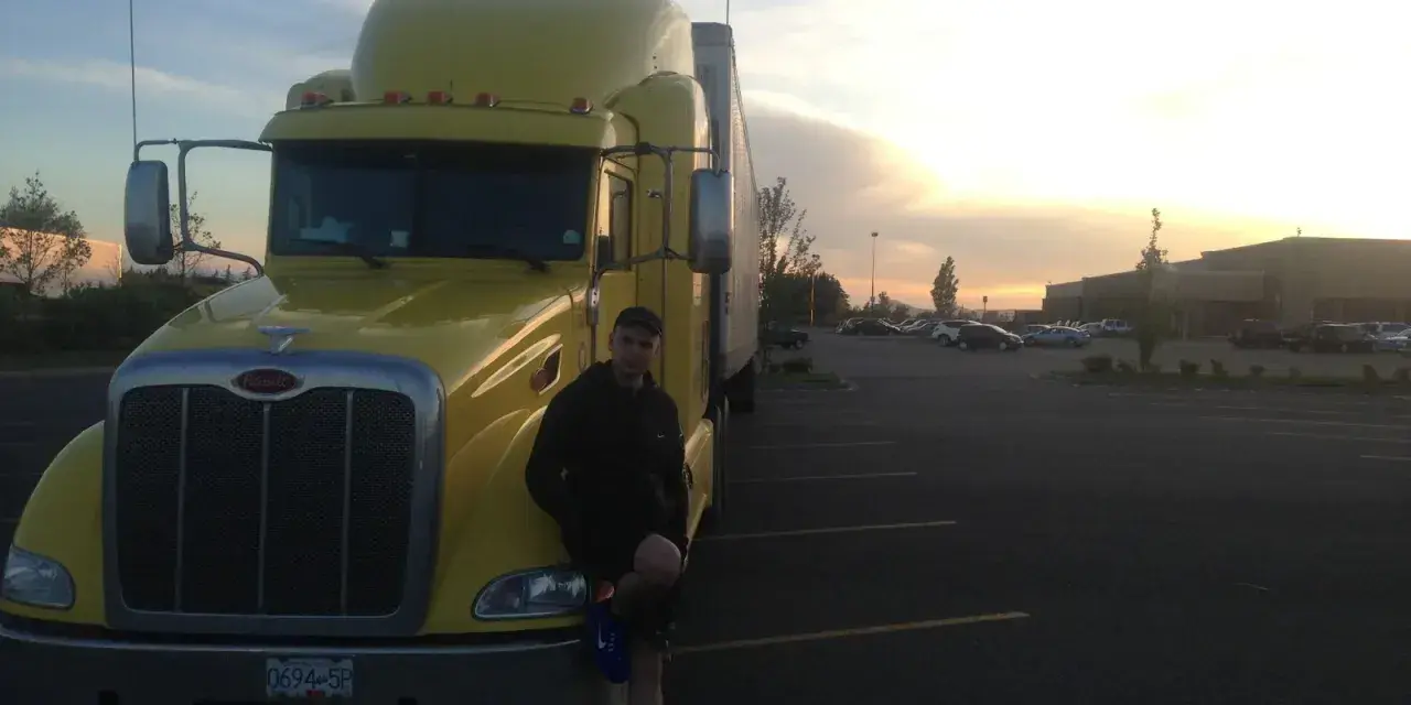 Młody mężczyzna opiera się o żółtą ciężarówkę. W tle zachód słońca i parking pełen aut. To obrazek z życia polskie truckerki.