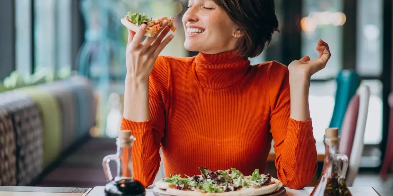 Kobieta w pomarańczowym golfie cieszy się pizzą, czując radość. W czym jest serotonina? W takich chwilach szczęścia!