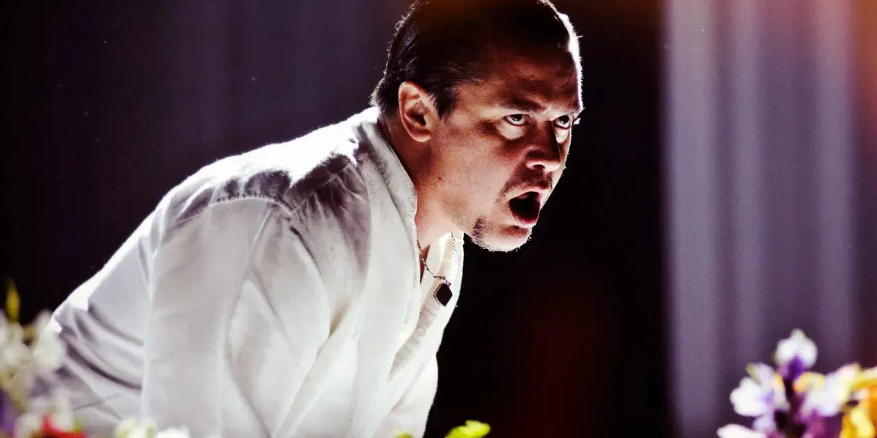 Mike Patton heute, mit weit aufgerissenem Mund und intensivem Blick, trägt ein weißes Hemd. Blumen im Vordergrund.