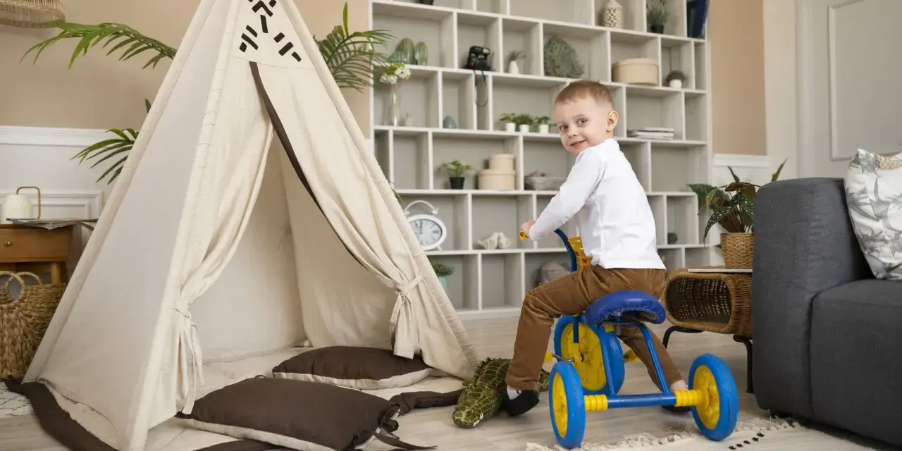 Un petit garçon sur un tricycle bleu et jaune s'amuse dans un espace de jeux, près d'une tente tipi et d'un canapé. Ce centre de vacances et de loisirs est idéal pour les enfants.