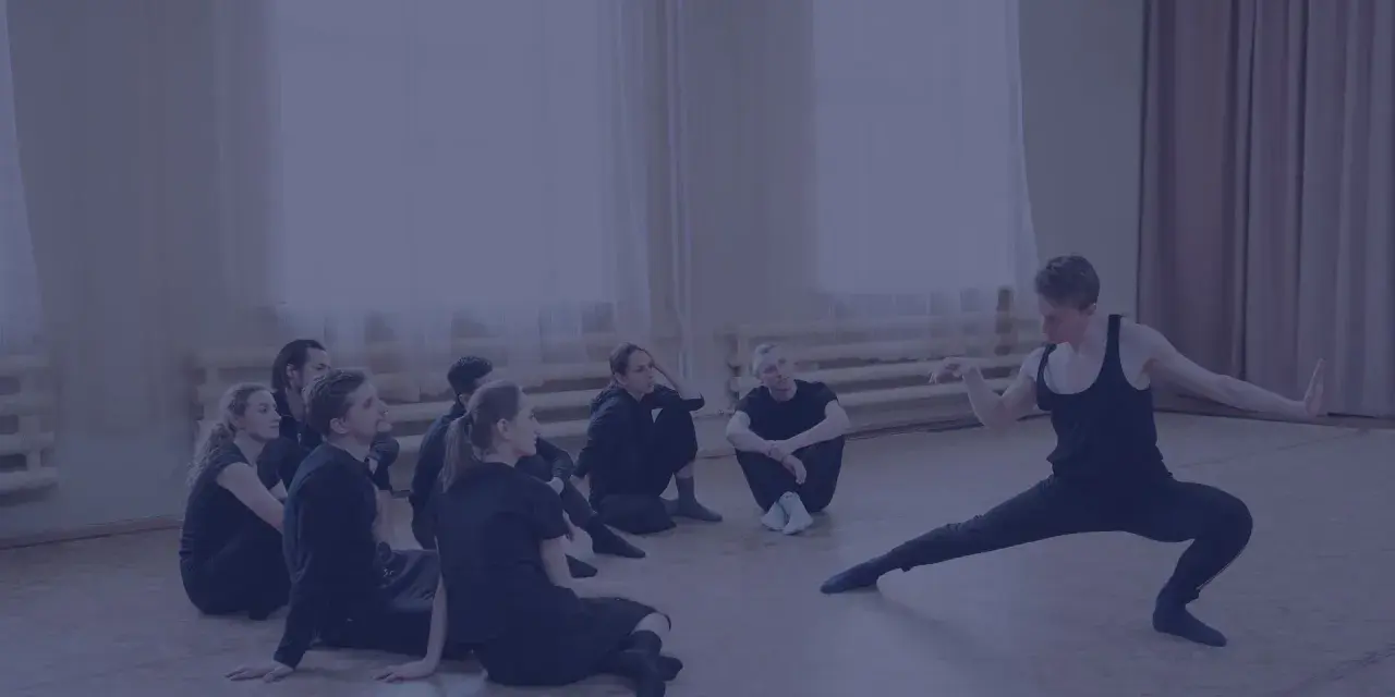 Choreograf demonstruje ruchy grupie tancerzy siedzących na podłodze.