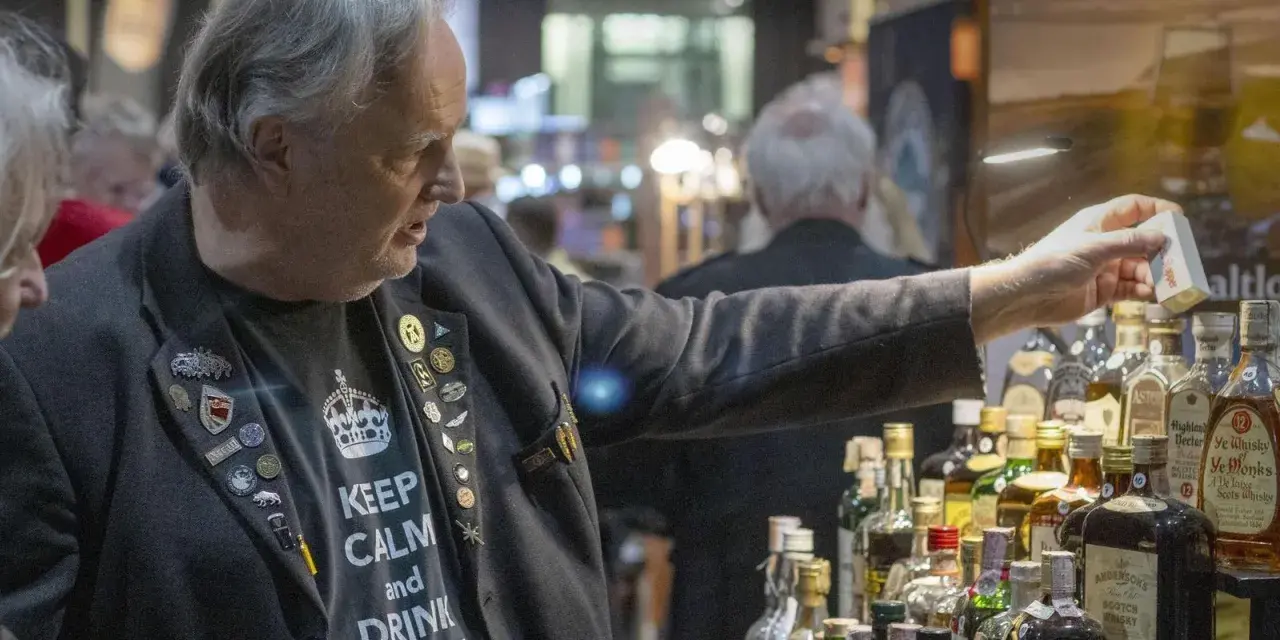 Mann mit "Keep Calm and Drink Whisky"-Shirt und vielen Ansteckern auf einer Whiskey Messe, der eine kleine Box hält.
