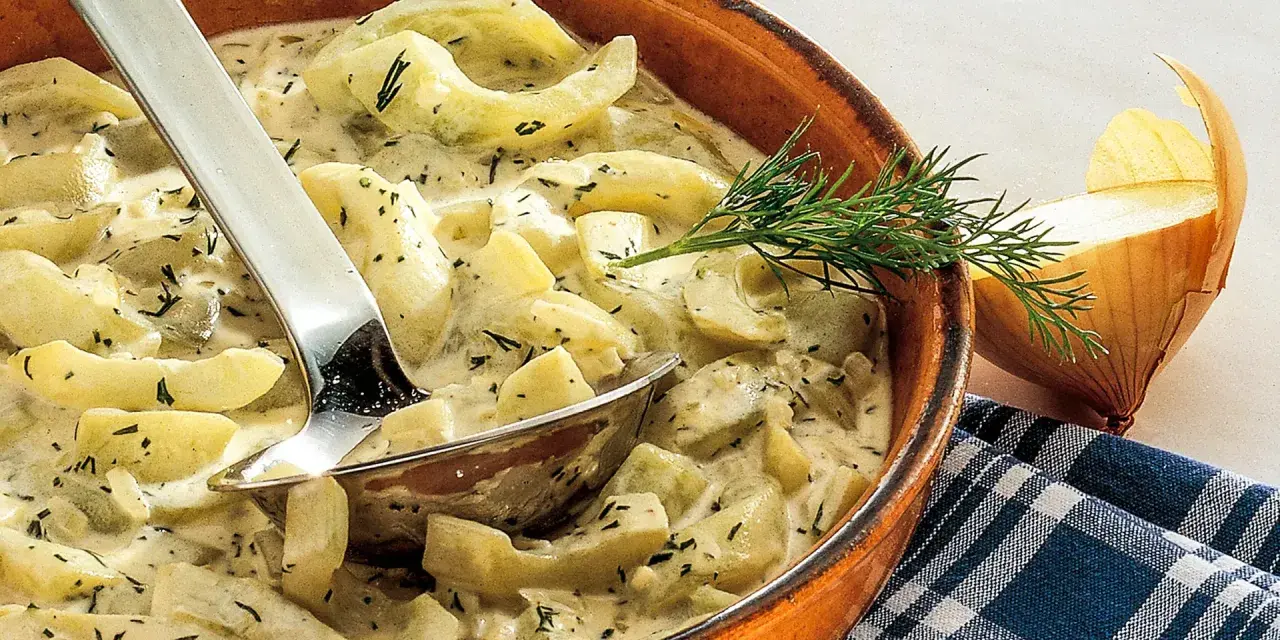 Cremiges Schmorgurken Rezept mit Dill in einer Tonschale, daneben eine Zwiebel und ein Löffel.