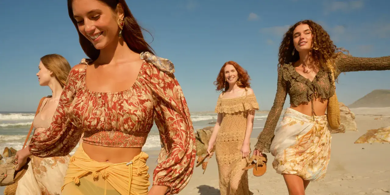 Quattro donne con abiti in stile boho passeggiano su una spiaggia sabbiosa con il mare sullo sfondo.