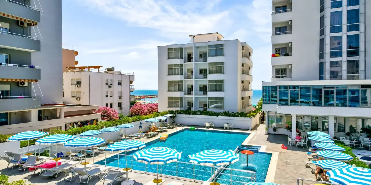 Albania - wczasy samolotem all inclusive. Basen hotelowy z leżakami i parasolami, w tle budynki i morze. Idealne miejsce na relaks.
