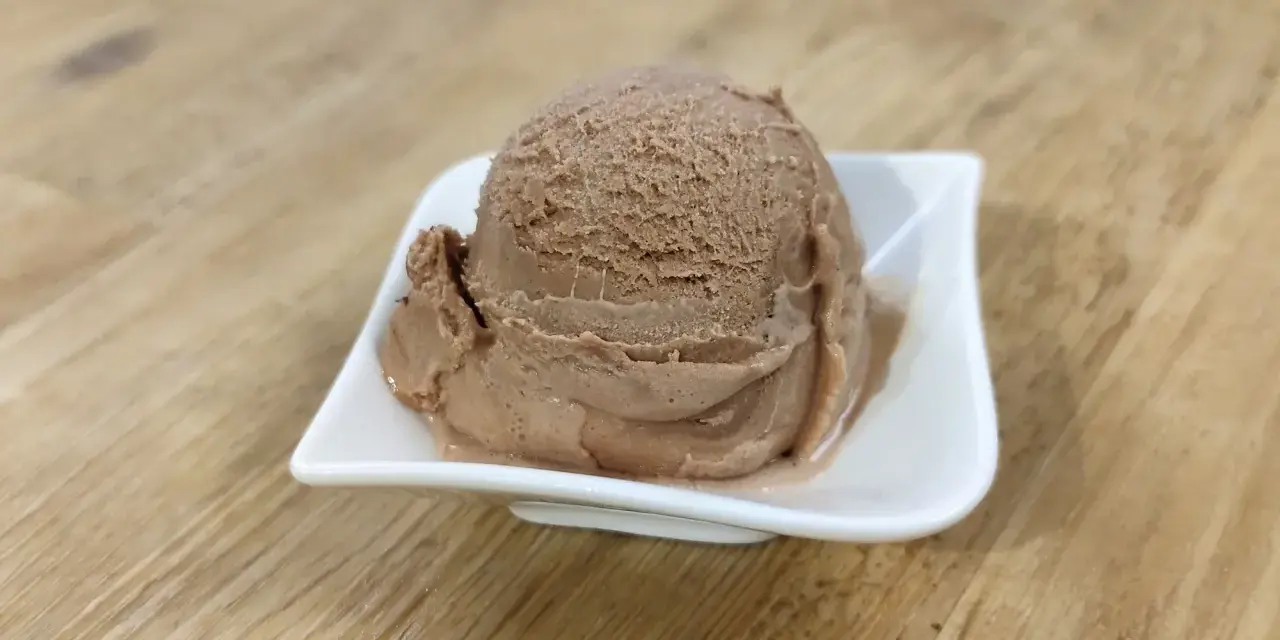 Ein Löffel cremiges Nougat Eis in einer weißen Schale auf einem Holztisch.