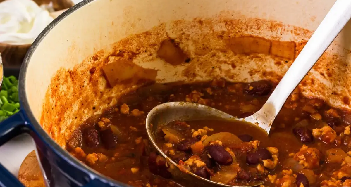 Gęste, aromatyczne chili con carne w garnku, z widoczną łyżką nabierającą porcję. Obok miseczka ze śmietaną.