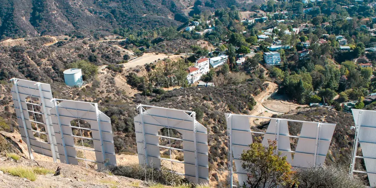 Widok zza gigantycznych liter znaku Hollywood. W oddali widać wzgórza z domami i drogami.