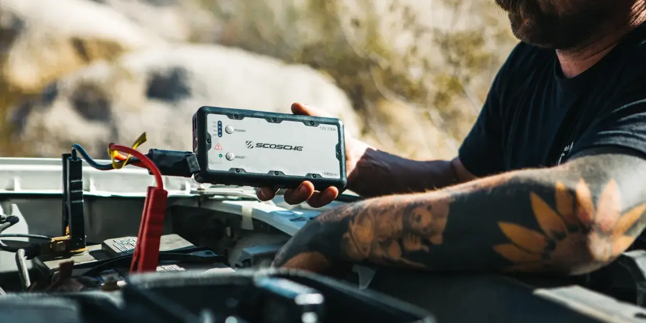 Man with tattoos demonstrates how to charge a jump starter by connecting it to a car battery in a desert landscape.