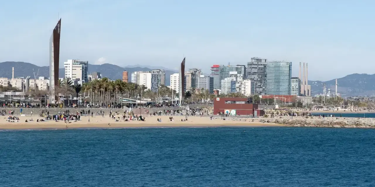 Ludzie odpoczywają na plaży, a w tle widać nowoczesne budynki Barcelony. To miasto leży nad Morzem Śródziemnym.