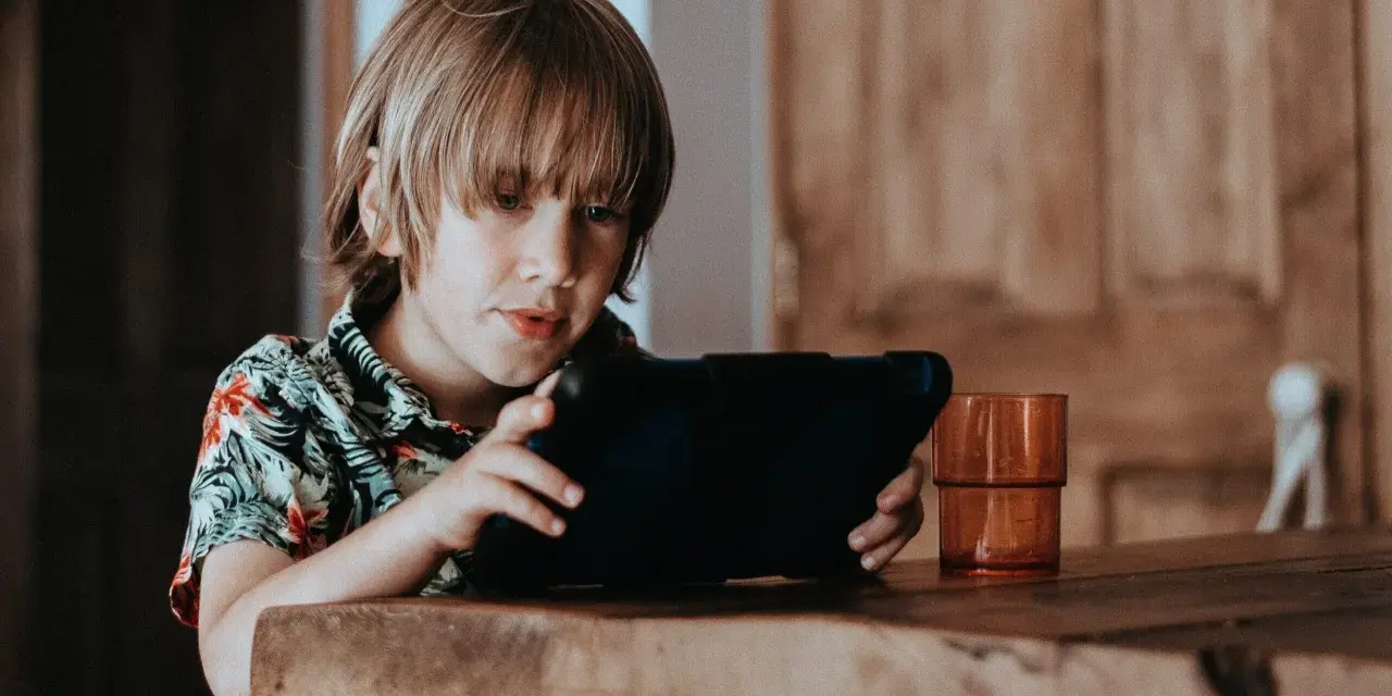 Chłopiec w hawajskiej koszuli ogląda krótkie filmy na tablecie, siedząc przy drewnianym stole.