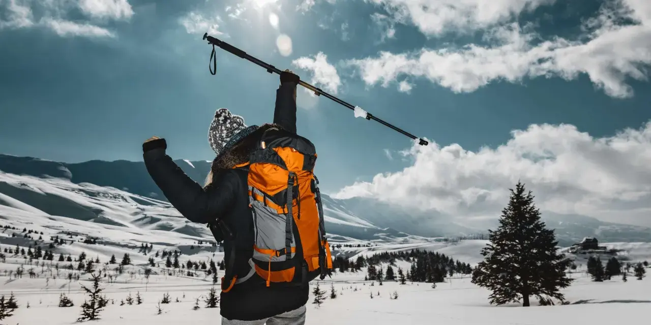 Osoba z plecakiem wznosi kijek, przygotowując się do marszu. Dowiedz się, jak prawidłowo założyć buciki na kijki nordic walking, by cieszyć się zimową wędrówką.