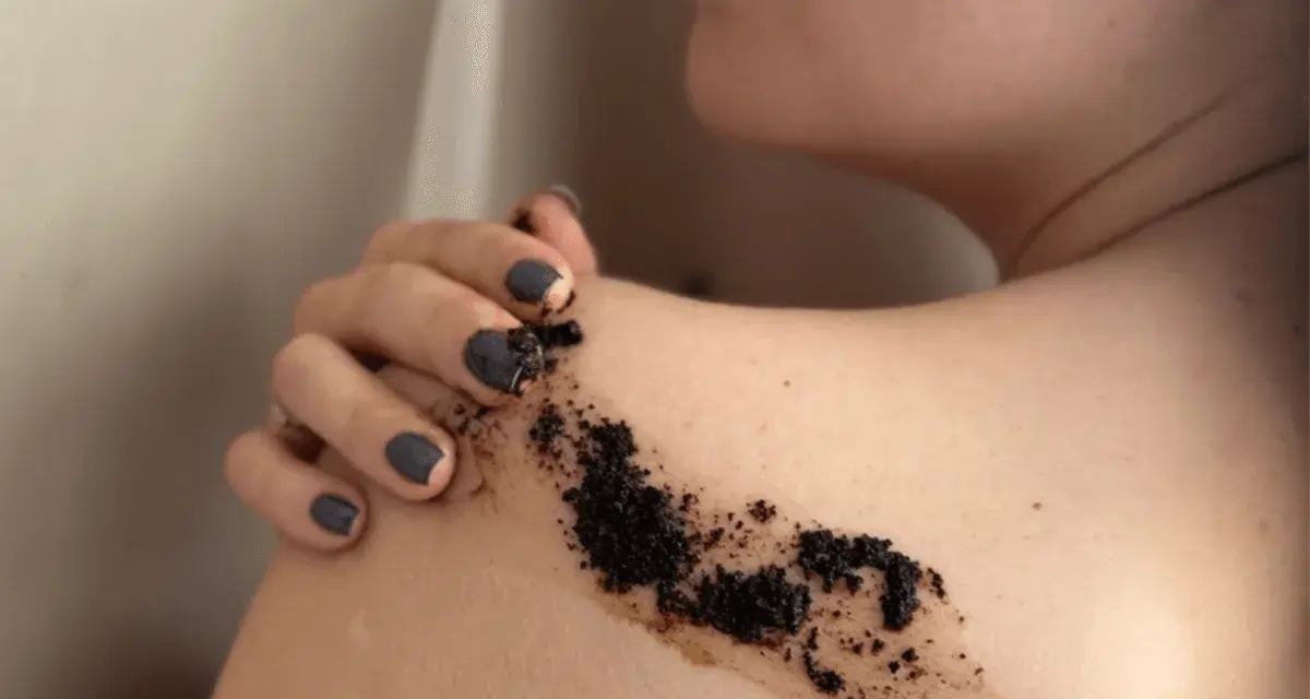 Dłoń z ciemnym lakierem nakłada peeling kawowy na ramię. Efekty po miesiącu regularnego stosowania są widoczne.