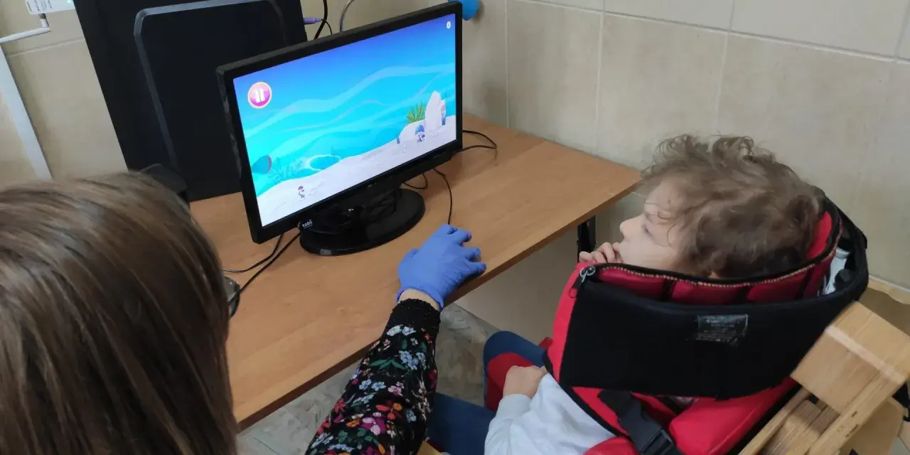 Tyflopedagog w niebieskiej rękawiczce pomaga dziecku w specjalnym foteliku grać w grę na komputerze.
