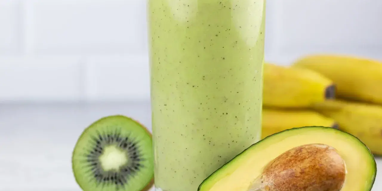 Zielony, kremowy koktajl z awokado, kiwi i banana. Obok szklanki połówka awokado i przekrojone kiwi. W tle pęk bananów.