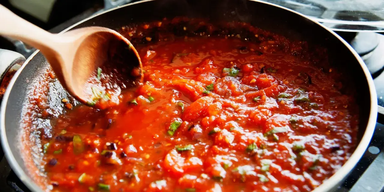 Gęsty, aromatyczny sos do spaghetti gotuje się na patelni. Drewniana łyżka miesza pomidorową masę z ziołami.