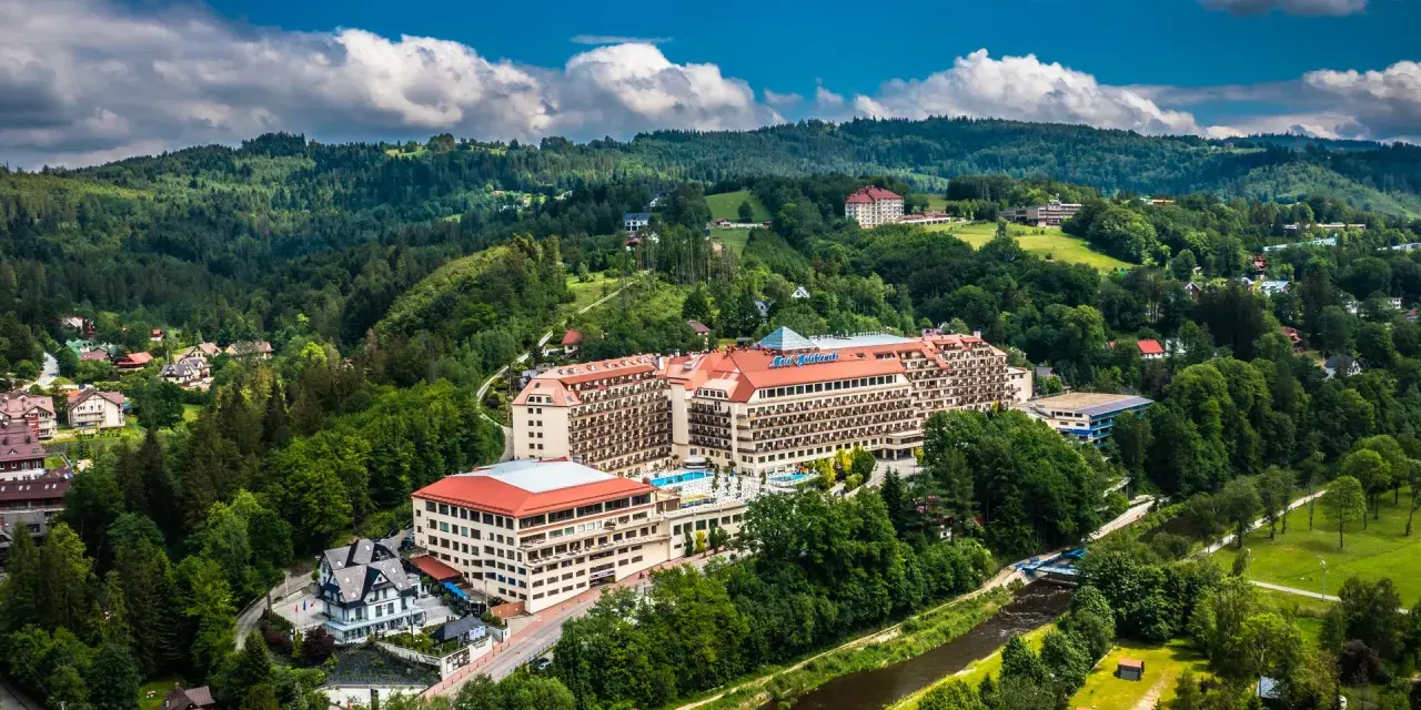 Widok z lotu ptaka na duży hotel Gołębiewski otoczony zielonymi wzgórzami i rzeką.