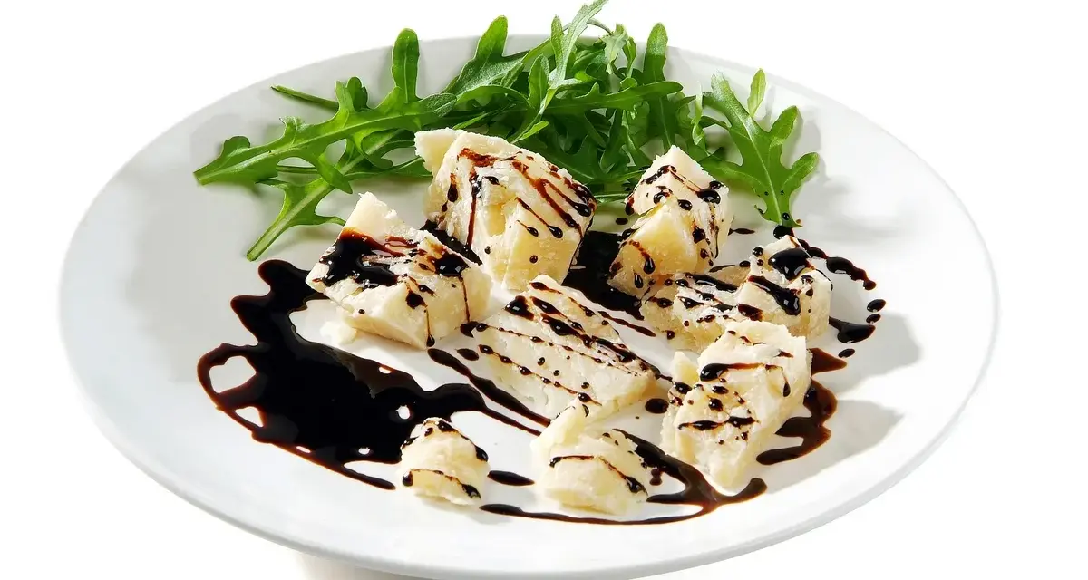 Kawałki parmezanu polane gęstą glazurą balsamiczną, podane z rukolą na białym talerzu.