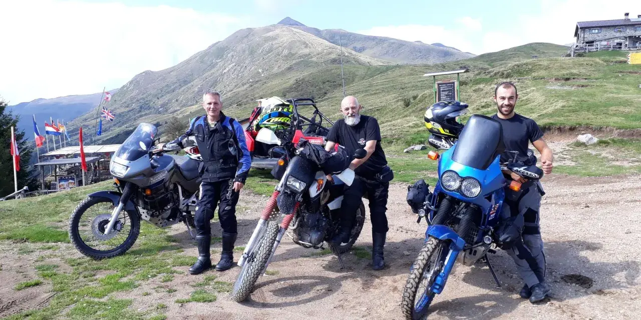 Tre motociclisti con moto da enduro posano in cima al passo San Lucio, con montagne sullo sfondo.
