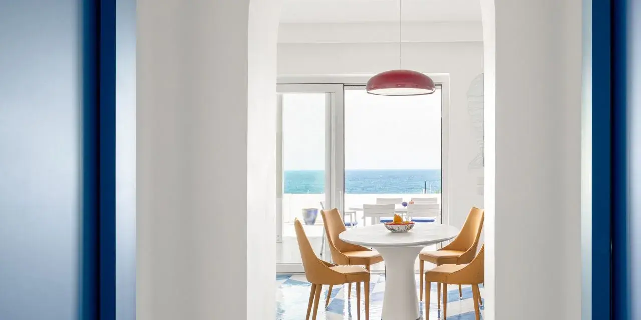 Interno di una casa stile mare con pavimento a scacchi blu e bianchi, tavolo rotondo con sedie arancioni e vista sul mare.