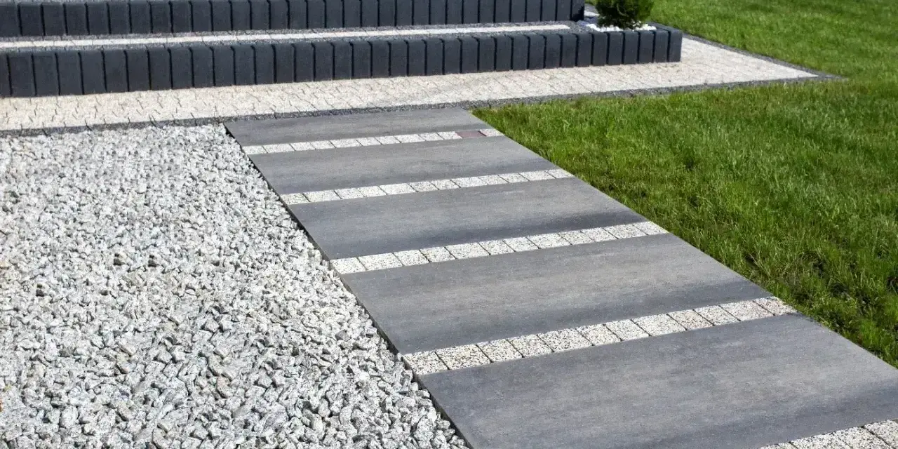 Nowy, tani chodnik przed domem z szarej płyty i białego gresu, obok żwiru i trawnika. Schody z ciemnego betonu.