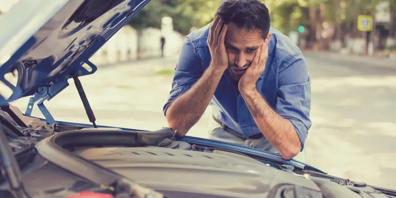 A frustrated man with hands on his head stares at his car's open hood, likely hearing a grinding noise when starting the car.
