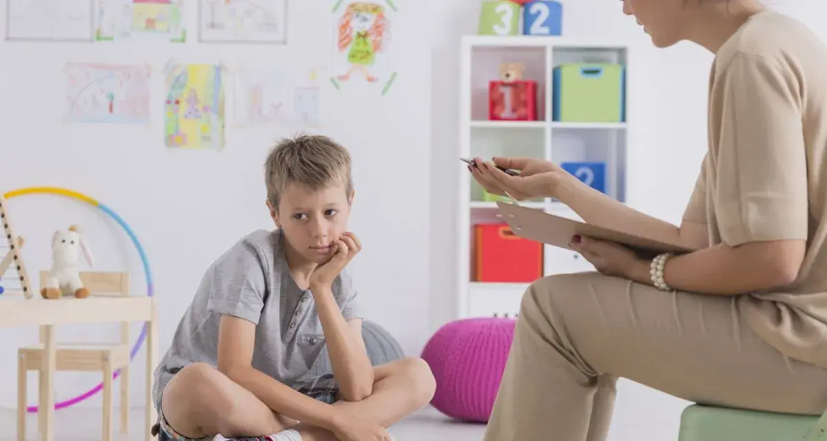 Chłopiec siedzi na poduszce, zastanawiając się, jakie pytania zadaje psycholog dziecku. W tle rysunki dzieci i kolorowe zabawki.