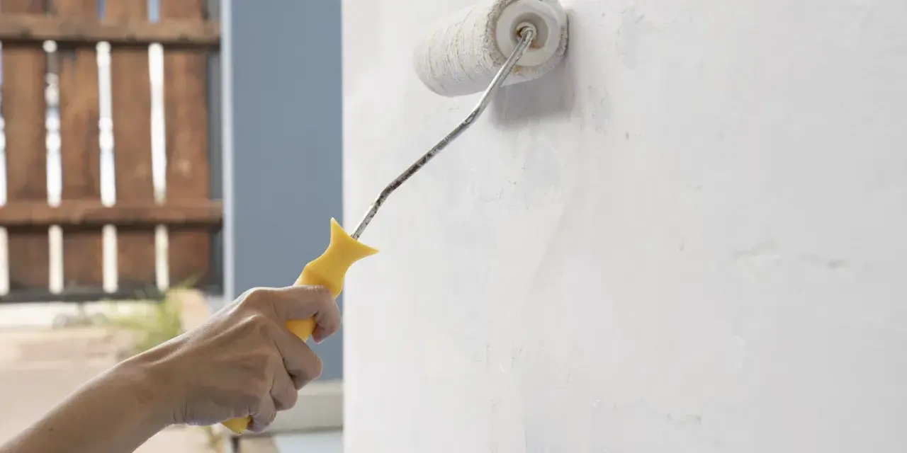 Mano aplica capa de imprimación blanca a una pared con un rodillo de pintura amarillo.
