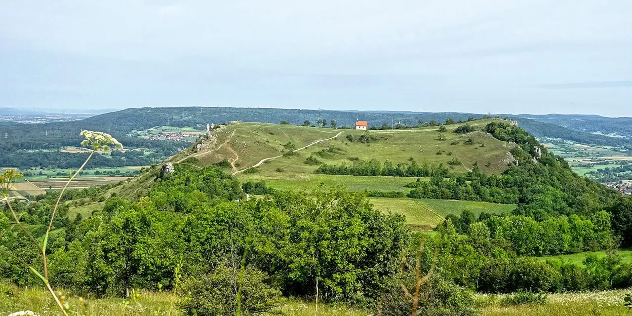Grüne Hügel und Felder laden zum Walberla wandern ein. Ein kleiner Weg schlängelt sich zum Gipfel mit einem roten Haus.