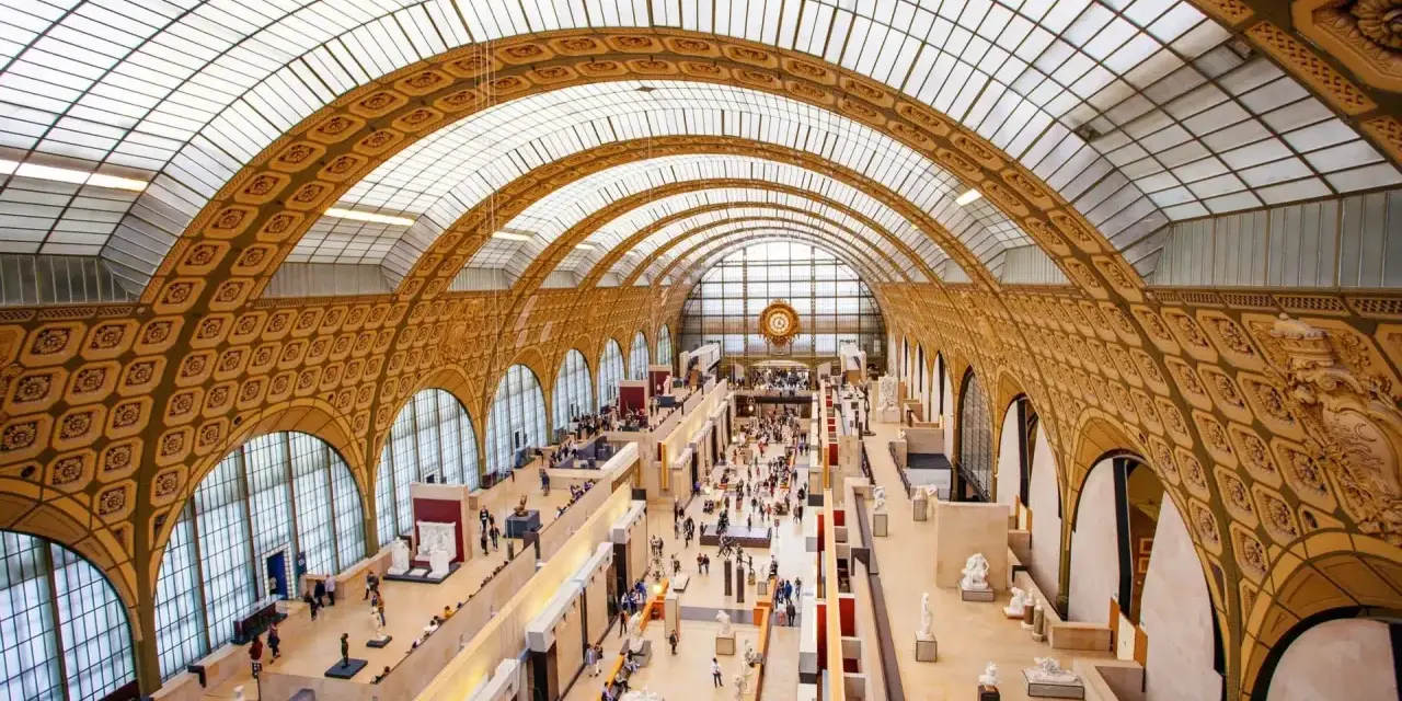 Interior del museo D'Orsay, con su impresionante techo abovedado de cristal y arcos dorados. Gente pasea entre esculturas y exposiciones.