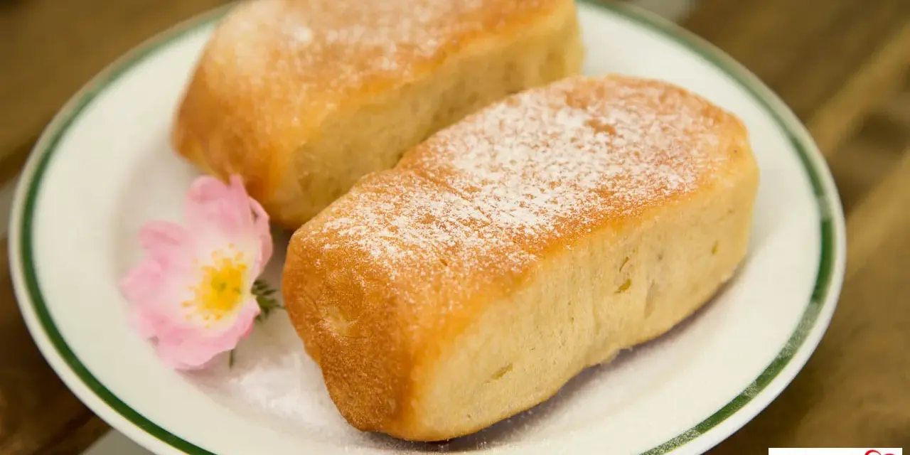 Zwei goldbraune Buchteln, bestäubt mit Puderzucker, auf einem Teller mit einer zarten rosa Blüte. Omas Rezept für Buchteln, ein Genuss!