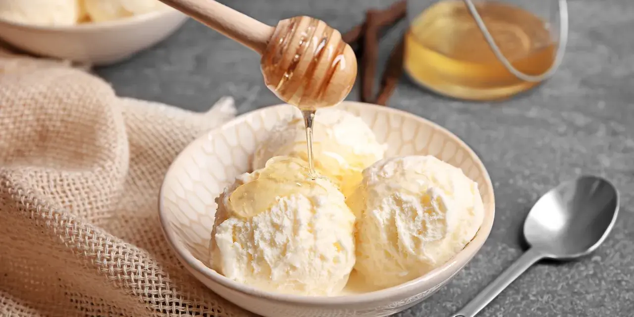Schneeballgroße Kugeln Vanilleeis werden mit flüssigem Honig beträufelt. Ein Holzlöffel tropft goldenen Honig auf das cremige Eis.