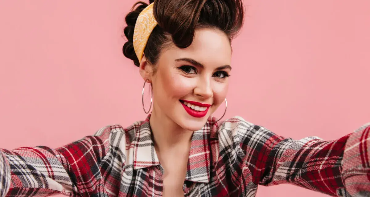 Uśmiechnięta kobieta w stylu pin-up, z burzą brązowych włosów spiętych w kok i żółtą bandaną, w kratę koszuli, robi sobie selfie.