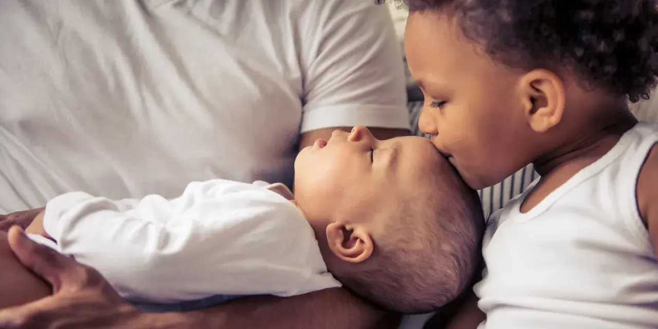 Ein älteres Kind küsst liebevoll die Stirn eines Babys. Die Frage "zweites Kind ja oder nein" scheint hier beantwortet.