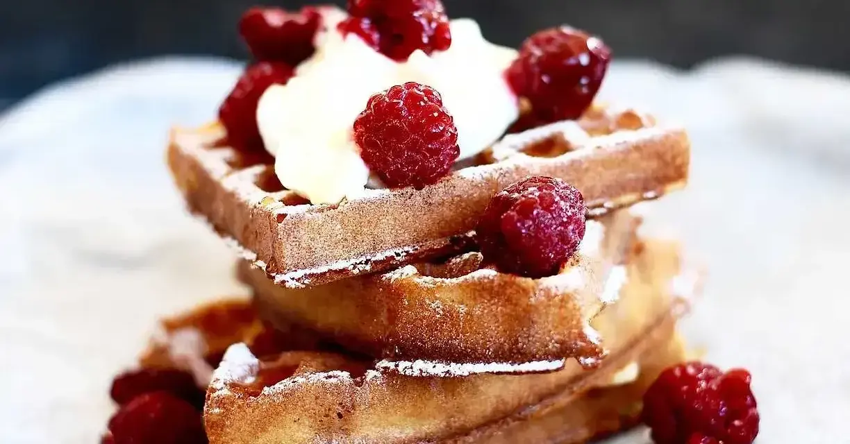 En stapel gyllene belgiska våfflor toppade med vispgrädde och färska hallon, pudrade med florsocker. En härlig frukost eller efterrätt.