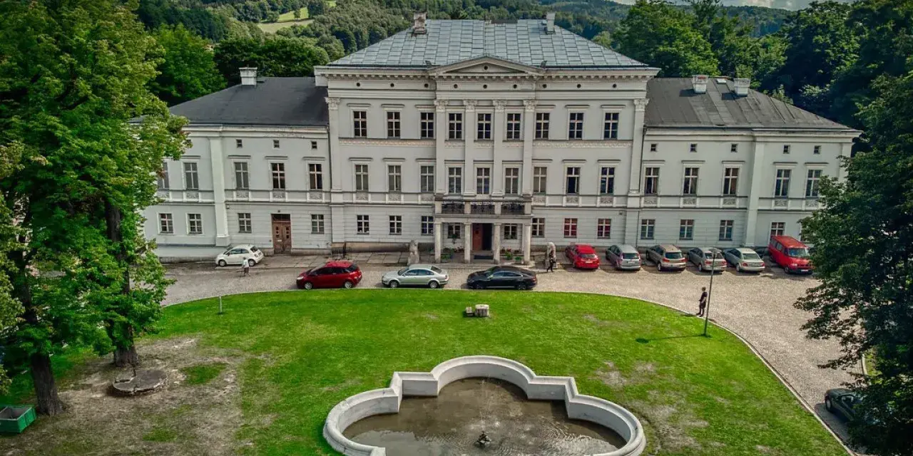 Pałac Jedlinka, biała, okazała budowla z szarym dachem, otoczona zielenią i górami. Przed pałacem fontanna i parking z samochodami.