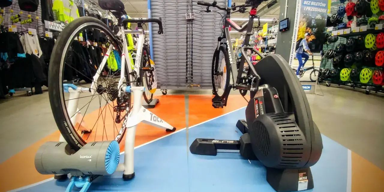Dwa rowery na trenażerach, jeden z napędem direct drive, gotowe do treningu w sklepie sportowym.