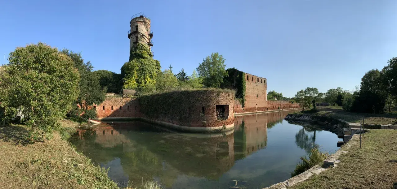 Antica torre e mura di mattoni circondate da un fossato, un angolo suggestivo di Venezia e dintorni, con vegetazione rigogliosa.