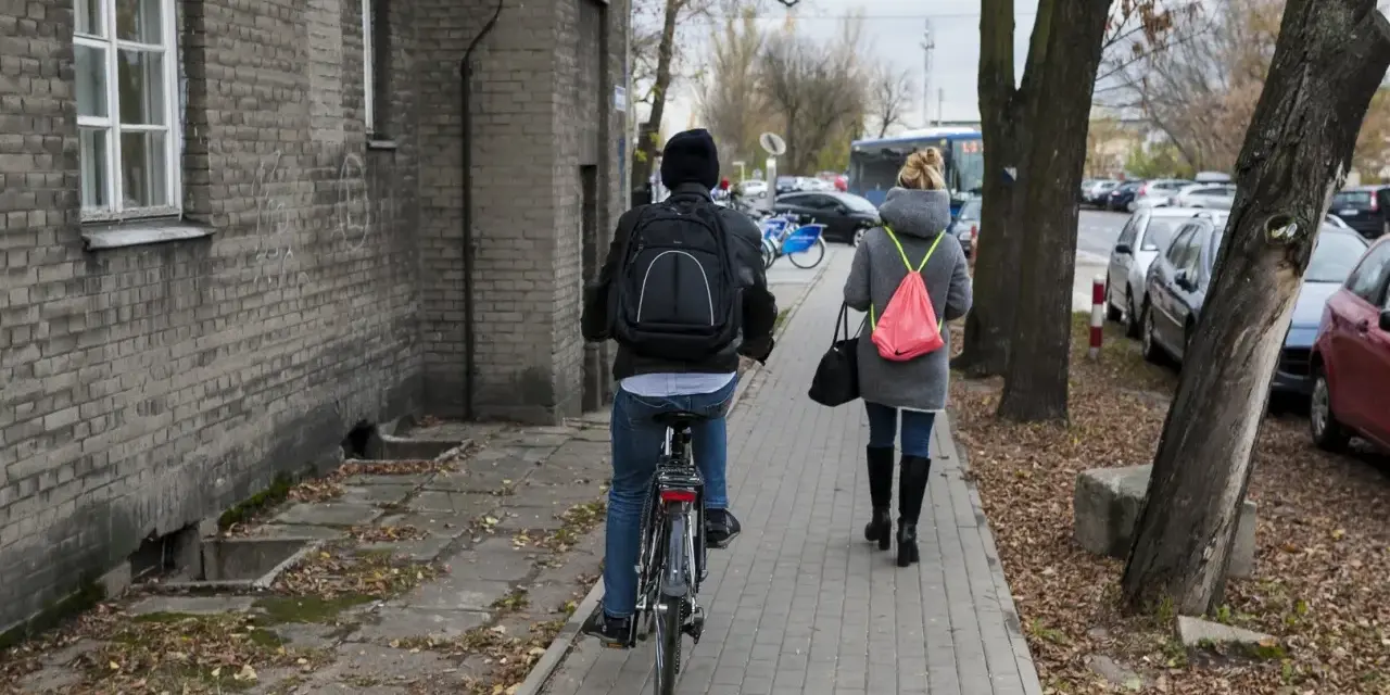 Osoba na rowerze i kobieta idąca chodnikiem obok budynku i drzew.