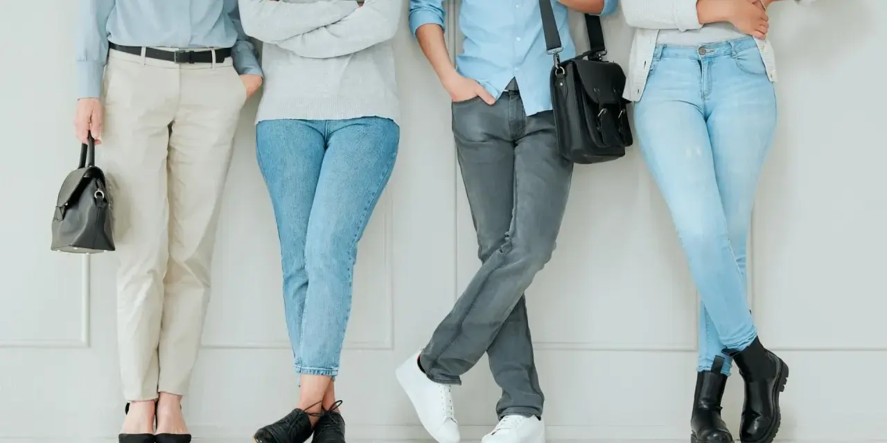 Cztery osoby w casual styl stoją przy ścianie. Jedna w jasnych spodniach i czarnych szpilkach, druga w jeansach i czarnych botkach, trzecia w szarych jeansach i białych sneakersach, czwarta w jasnych jeansach i czarnych półbutach.