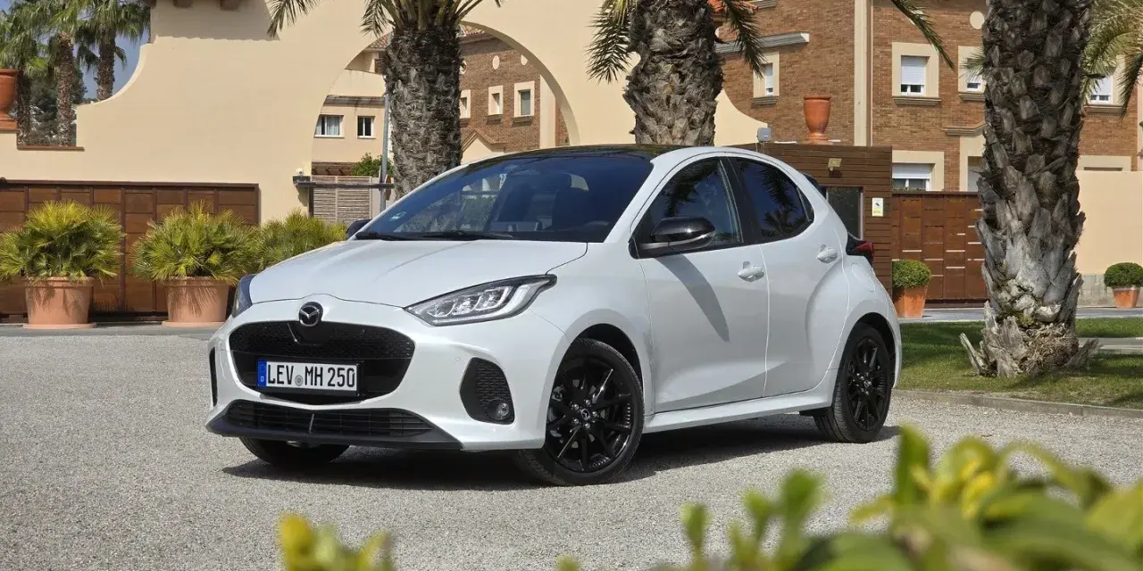 Biała Mazda 2 Hybrid zaparkowana przed budynkiem z palmami. Stylowy miejski samochód gotowy na podróż.