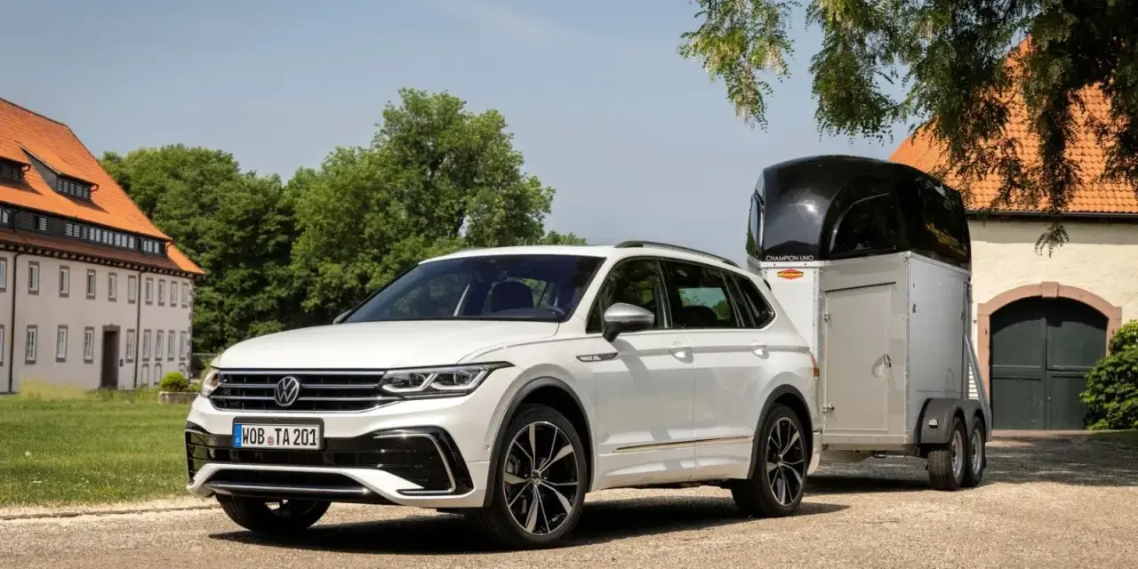 Biały Volkswagen SUV z przyczepą do przewozu koni stoi przed starym budynkiem.