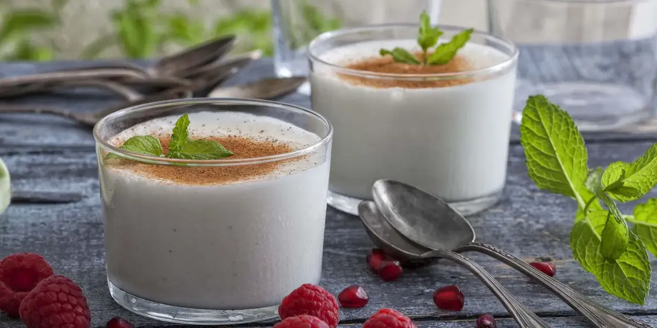 Zwei Gläser cremiger Leche Merengada, garniert mit Minze und Zimt, umgeben von Himbeeren und Granatapfelkernen.