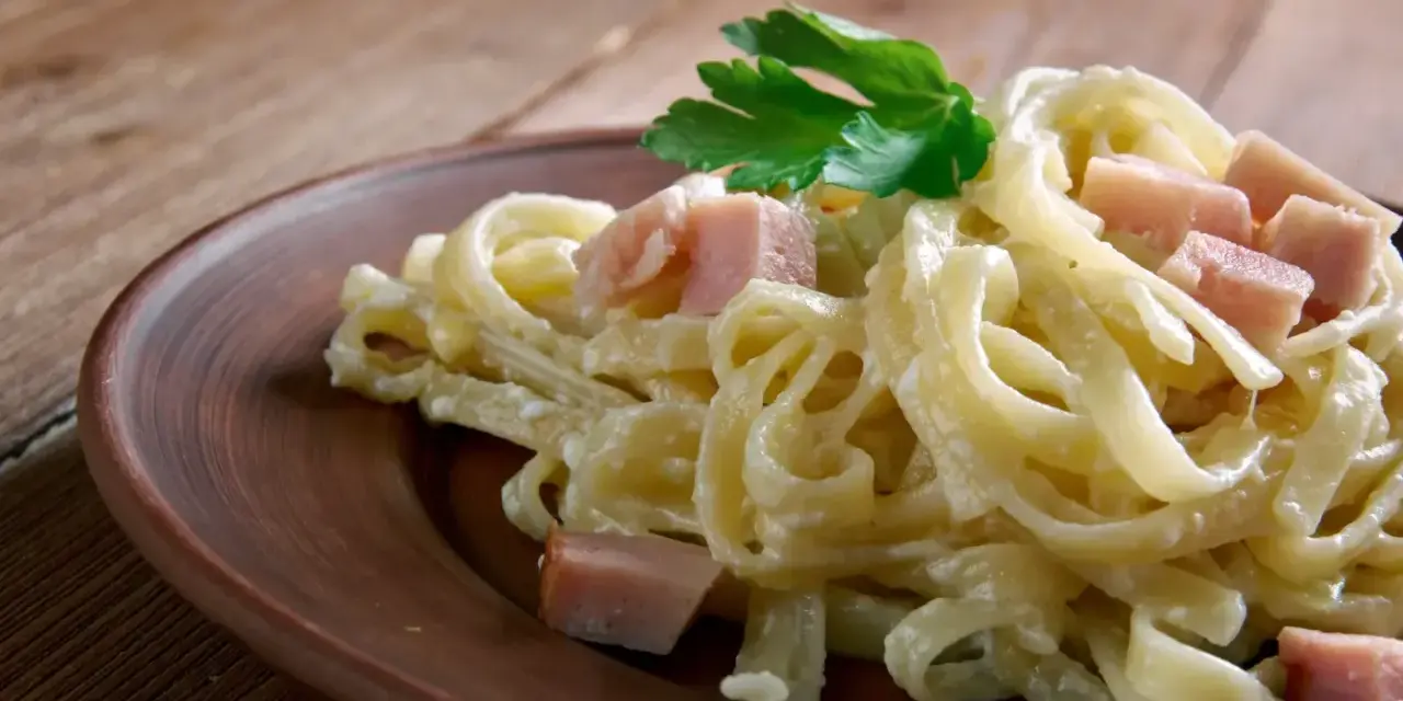 Fettuccine co to? Pyszne, kremowe fettuccine z kawałkami szynki i natką pietruszki na brązowym talerzu.